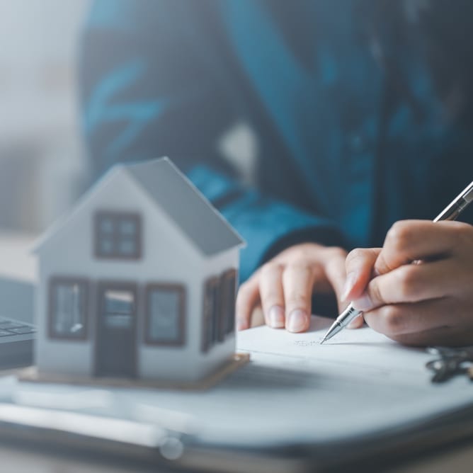 A hand signing paperwork on a desk beside a model house, a laptop and a set of keys