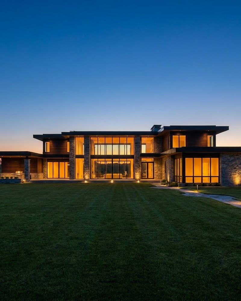 A large modern stone and timber house at dusk with its full-height windows lit warm, seen across a mown lawn