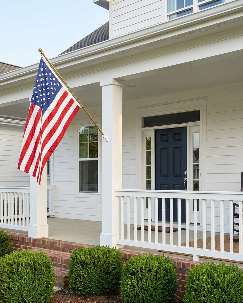 A United States flag on a brass pole beside the white covered porch and navy front door of a home