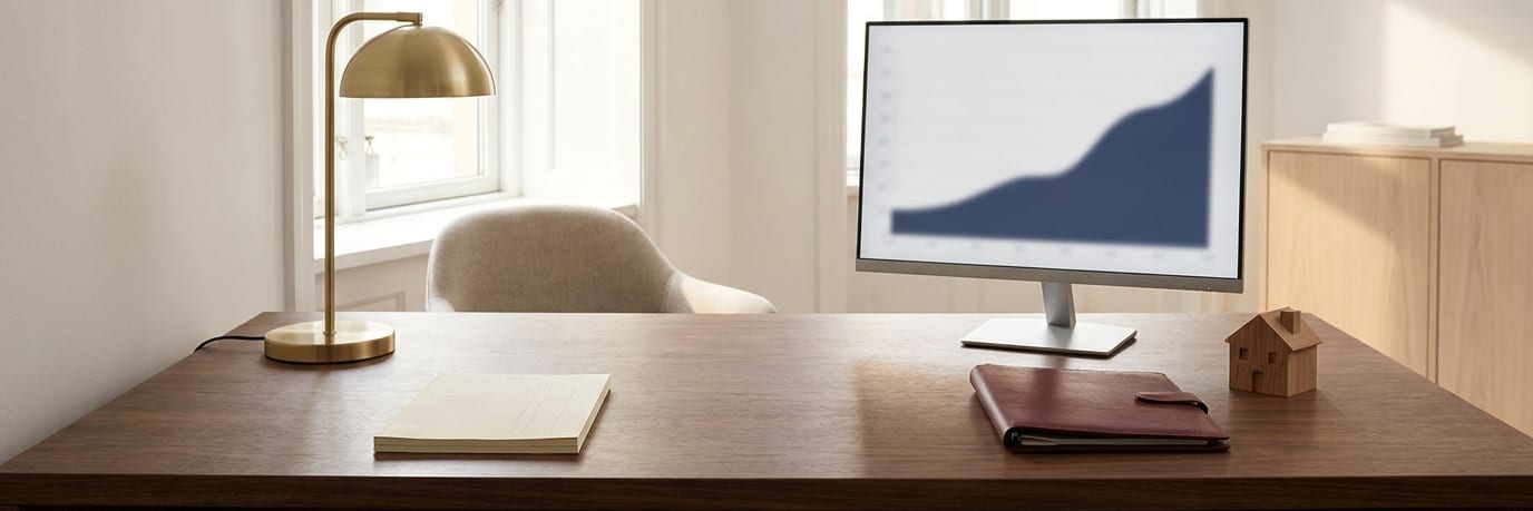 A walnut office desk with a monitor turned toward the visitor’s chair showing a rising navy chart, a brass lamp, a leather folder and a small wooden model house